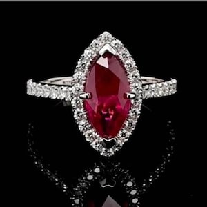 Può includere: Un anello con rubino a taglio marquise, circondato da un'aureola di piccoli diamanti. La fascia dell'anello è anch'essa incastonata con diamanti, in argento o oro bianco. L'anello è esposto su una superficie nera, che ne esalta la brillantezza.