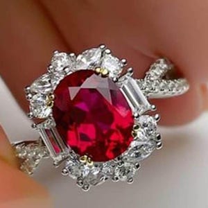 Puede incluir: Un anillo de plata con una gran piedra preciosa de rubí de corte ovalado rodeada de piedras preciosas más pequeñas y transparentes. El rubí es de color rojo intenso y el anillo tiene un diseño detallado y ornamentado. El anillo está engastado con varias piedras preciosas pequeñas y transparentes.