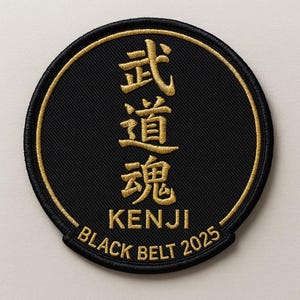 May include: A circular black patch with gold embroidery. It features Japanese characters, the name "KENJI", and the text "BLACK BELT 2025". The patch is likely for martial arts or a similar discipline.