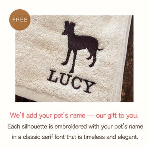 Puede incluir: Una toalla beige con una silueta de perro marr&oacute;n oscuro y el nombre "LUCY" bordado en una fuente serif cl&aacute;sica. Una etiqueta "FREE" est&aacute; en la esquina superior izquierda. La toalla est&aacute; doblada y parece estar hecha de un material suave y absorbente.