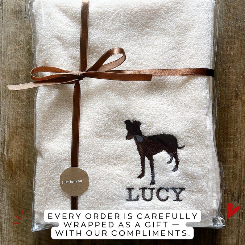Puede incluir: Una toalla blanca bordada con la silueta de un perro negro y el nombre "LUCY" en letras negras. La toalla est&aacute; envuelta en pl&aacute;stico transparente y atada con una cinta marr&oacute;n. Una peque&ntilde;a etiqueta dice "Just for you".