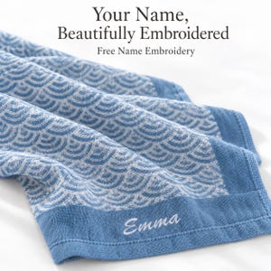 Op de afbeelding: Een blauwe en witte handdoek met een herhalend golfpatroon en een effen blauwe rand. De naam Emma is in wit schrift geborduurd op de onderrand. De tekst boven de handdoek luidt: Your Name, Beautifully Embroidered, Free Name Embroidery.
