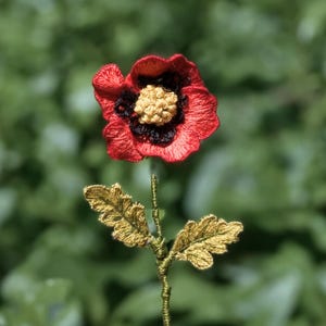 May include: A handcrafted, embroidered flower with a vibrant red bloom, a black center, and a yellow stamen. The stem and leaves are a golden-yellow color, set against a blurred green background. This floral art piece is a unique decorative item.