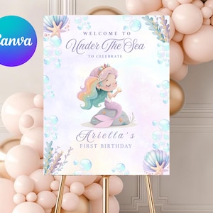 May include: A sign with the text "Welcome to Under The Sea to Celebrate" featuring a mermaid illustration. The sign is for Ariella's first birthday. The Canva logo is in the upper left corner. The sign is on a gold easel.