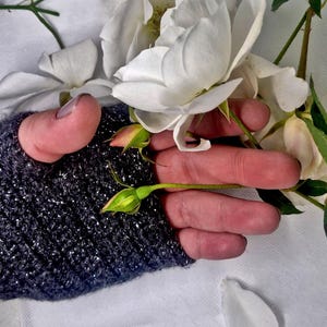 K&ouml;nnte beinhalten: Nahaufnahme von wei&szlig;en Rosen und Knospen. Die Blumen werden von einer Hand gehalten, die einen dunkelgrauen, glitzernden Fingerhandschuh tr&auml;gt. Der Hintergrund ist ein weiches wei&szlig;es Tuch mit ein paar verstreuten Bl&uuml;tenbl&auml;ttern.