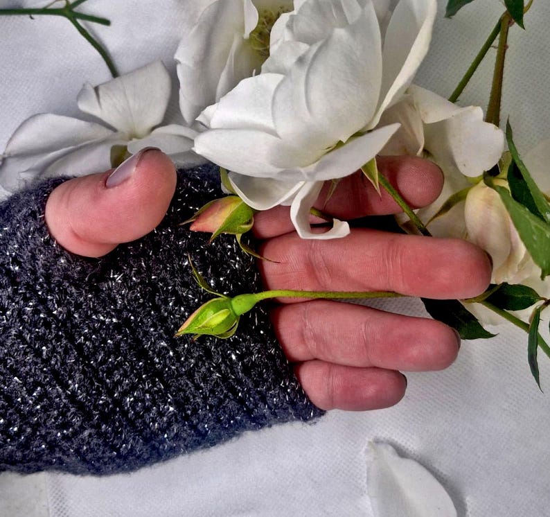 K&ouml;nnte beinhalten: Nahaufnahme von wei&szlig;en Rosen und Knospen. Die Blumen werden von einer Hand gehalten, die einen dunkelgrauen, glitzernden Fingerhandschuh tr&auml;gt. Der Hintergrund ist ein weiches wei&szlig;es Tuch mit ein paar verstreuten Bl&uuml;tenbl&auml;ttern.