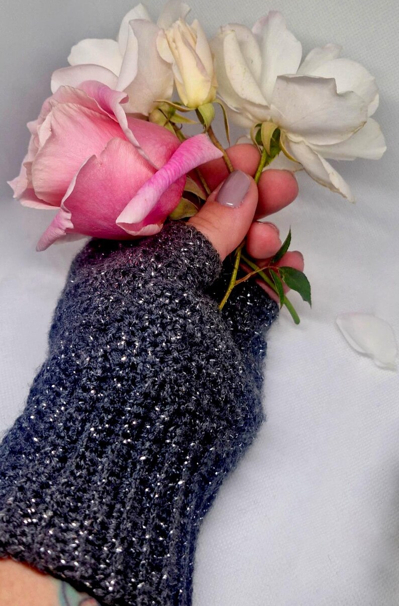 K&ouml;nnte beinhalten: Ein dunkelgrauer fingerloser Handschuh mit silbernen Sprenkeln, der einen Strau&szlig; rosa und wei&szlig;er Rosen h&auml;lt. Der gestrickte Handschuh hat ein strukturiertes Aussehen, und die Rosen sind in voller Bl&uuml;te.
