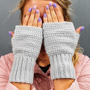 K&ouml;nnte beinhalten: Ein Paar hellgraue, fingerlose H&auml;kelhandschuhe. Die Handschuhe haben eine gerippte Manschette und ein strukturiertes Stichmuster am Handteil. Das Bild zeigt die Handschuhe, die hochgehalten werden und das Gesicht bedecken.