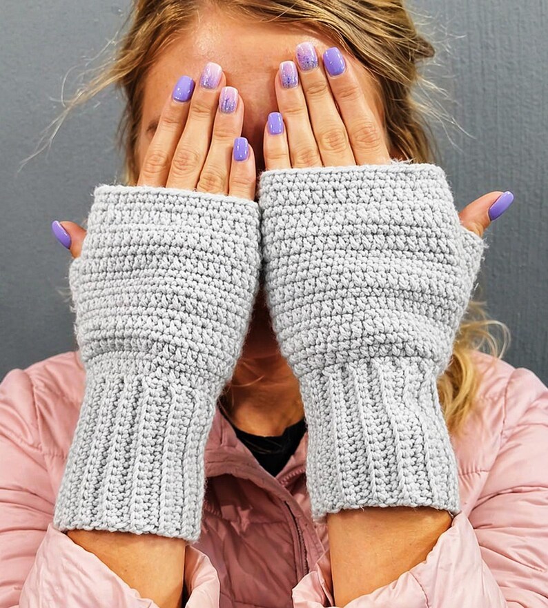 K&ouml;nnte beinhalten: Ein Paar hellgraue, fingerlose H&auml;kelhandschuhe. Die Handschuhe haben eine gerippte Manschette und ein strukturiertes Stichmuster am Handteil. Das Bild zeigt die Handschuhe, die hochgehalten werden und das Gesicht bedecken.