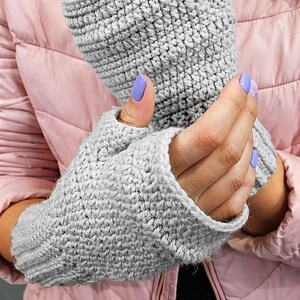 K&ouml;nnte beinhalten: Ein Paar graue Fingerhandschuhe, handgeh&auml;kelt mit einem strukturierten Stich. Die Handschuhe sind aus einem weichen, hellgrauen Garn gefertigt. Die Handschuhe werden an einer Person gezeigt, die eine rosa Jacke tr&auml;gt.