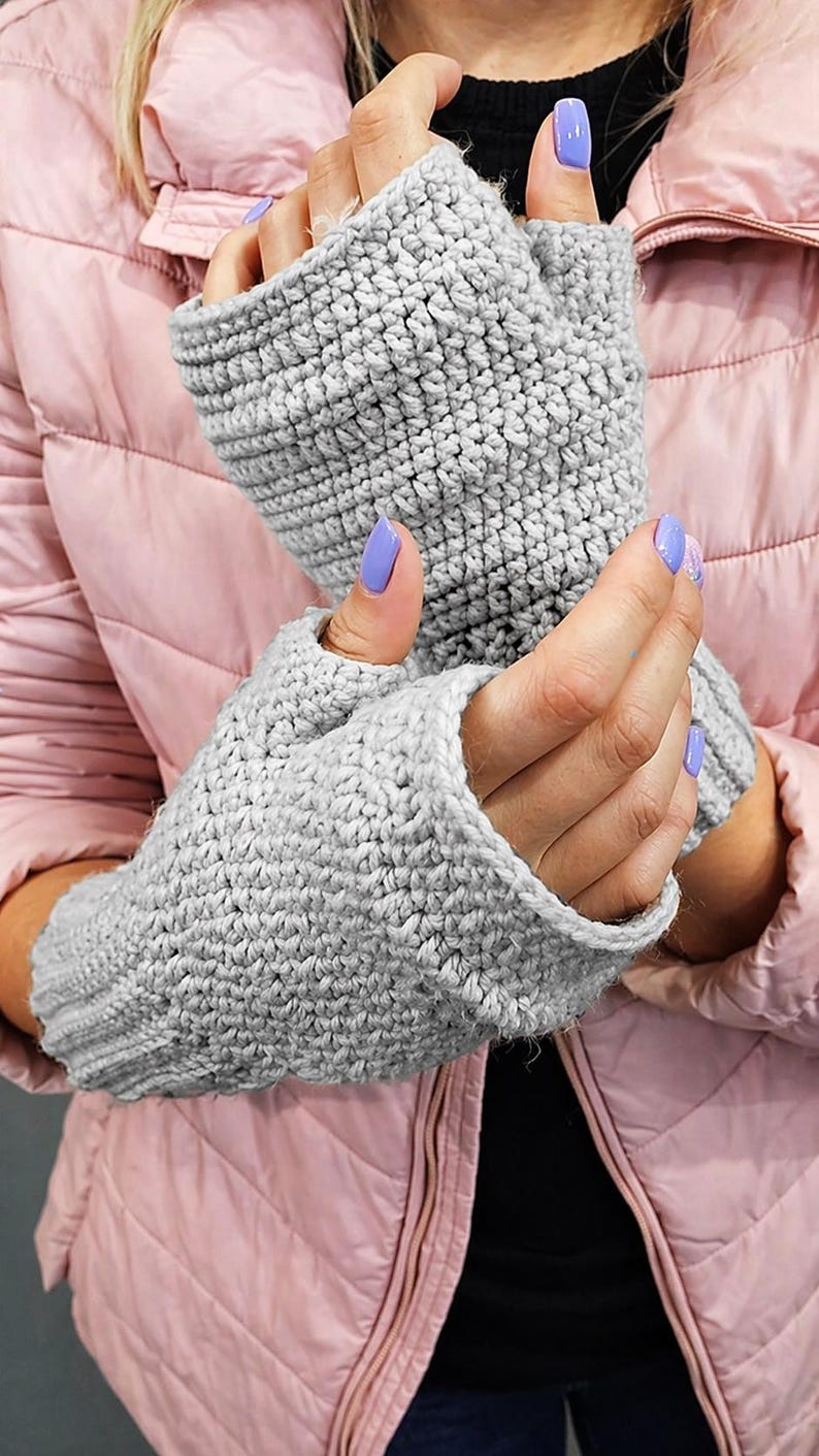 K&ouml;nnte beinhalten: Ein Paar graue Fingerhandschuhe, handgeh&auml;kelt mit einem strukturierten Stich. Die Handschuhe sind aus einem weichen, hellgrauen Garn gefertigt. Die Handschuhe werden an einer Person gezeigt, die eine rosa Jacke tr&auml;gt.