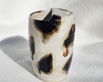 May include: A ceramic tumbler with an off-white base glaze featuring irregular, dark brown and black splotches that resemble organic spots. The vessel has a slightly uneven, hand-formed rim and a smooth, glossy finish across the exterior surface.