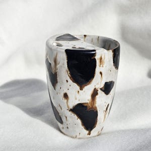 Könnte beinhalten: Ein Keramikbecher mit weißer Grundfarbe und unregelmäßigen schwarzen sowie braunen Flecken. Das Gefäß hat eine konisch zulaufende zylindrische Form mit einem markanten Randdesign, das an einer Seite eine kleine Aussparung aufweist.
