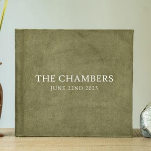 Peut inclure: Un album photo carré vert sauge avec le texte "THE CHAMBERS" et "JUNE 22ND 2025" en blanc. L'album est sur une surface en bois, à côté d'un vase décoratif avec de grandes plantes vertes et une géode.