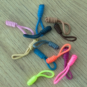 Könnte beinhalten: Eine Sammlung farbenfroher Reißverschluss-Anhänger in verschiedenen Farbtönen, darunter Rosa, Blau, Orange und Grün. Jeder Anhänger hat eine Schlaufenschnur und einen strukturierten, zylindrischen Griff. Die Anhänger sind auf einer hellbraunen Holzoberfläche angeordnet.