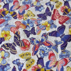 Op de afbeelding: Een stofpatroon met een dichte rangschikking van vlinders in tinten rood, blauw, geel en paars, afgewisseld met kleine bloemen en delicaat lijngetekend gebladerte op een witte achtergrond.