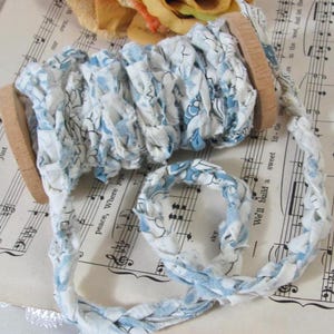 May include: A spool of light blue and white floral patterned fabric ribbon wrapped around a wooden bobbin. The ribbon is made of strips of fabric that have been braided together to create a textured, decorative trim with a soft, frayed appearance.