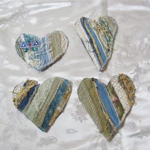 May include: Four heart-shaped fabric ornaments constructed from layered strips of blue denim, green fabric, white lace, and gold-toned trim. One heart features a blue and green butterfly-shaped embellishment with small rhinestones attached to the surface.