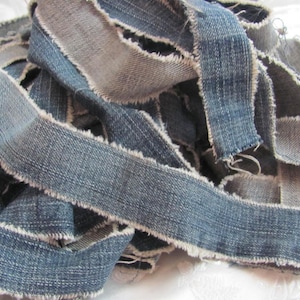 Pode incluir: Close-up de uma pilha de tiras de ganga em vários tons de azul e cinza. As tiras têm bordas desfiadas, criando uma aparência texturizada e rústica. Ideal para projetos de artesanato ou upcycling.