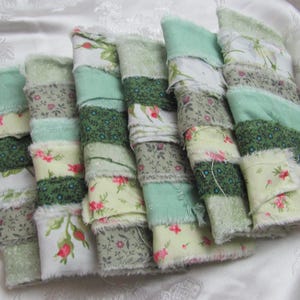 May include: A collection of fabric strips in shades of green, cream, and floral patterns. The strips are layered and stitched together, with frayed edges. The overall aesthetic is rustic and handmade, suggesting a craft project or decorative item.