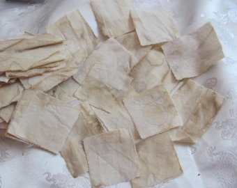 May include: A collection of small, square fabric patches in varying shades of beige and brown. The patches appear to be handmade, with visible edges and subtle staining, suggesting a vintage or rustic aesthetic. They are arranged on a white, textured surface.