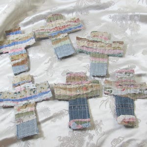 May include: Five cross-shaped ornaments constructed from various strips of fabric, including denim and floral patterns. Each cross features visible stitching and frayed edges, showcasing a patchwork design with a mix of blue, pink, and neutral tones.