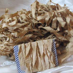 May include: A pile of tan fabric strips with frayed edges. A smaller piece of the same fabric is folded and has a blue and white patterned border. The fabric appears to be cotton or linen.