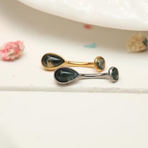 Pode incluir: Duas peças de joalheria decorativas. Uma é dourada, a outra prateada. Cada uma apresenta uma pedra verde em forma de gota e uma pedra redonda verde menor. As joias são exibidas em uma superfície branca com detalhes florais.