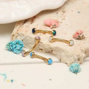 Puede incluir: Varios piercings de ceja curvos dorados, cada uno con una gema opalescente diferente. Las joyas se exhiben sobre una superficie clara y texturizada, con pequeñas flores secas esparcidas. Las gemas son de color azul, blanco e iridiscente.