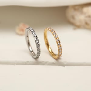 Op de afbeelding: Twee ringen, één zilverkleurig en één goudkleurig, elk versierd met een rij kleine, heldere edelstenen. De ringen staan tegen een zachte, neutrale achtergrond, wat hun delicate ontwerp en glans benadrukt. De gouden ring heeft een warme, stralende tint.