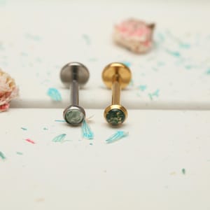 Puede incluir: Dos piercings para labios. Uno es de color plateado, el otro dorado. Ambos tienen un disco plano en la parte superior y una pequeña piedra redonda verde en la parte inferior. Los piercings están sobre una superficie blanca con confeti azul y rosa esparcido.