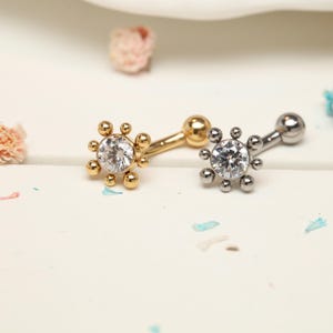 Könnte beinhalten: Zwei Bauchnabelpiercings. Eines ist goldfarben mit einem klaren, runden Mittelstein und kleinen goldenen Perlen darum. Das andere ist silberfarben mit einem klaren, runden Mittelstein und kleinen silbernen Perlen. Beide haben ein gebogenes Barbell-Design.