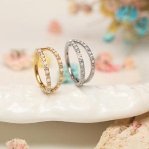 Peut inclure: Deux boucles d'oreilles doubles, l'une dorée et l'autre argentée, sont présentées sur une surface blanche. Les deux boucles d'oreilles comportent une rangée de petites pierres blanches et de cristaux transparents. Elles sont conçues pour les piercings.
