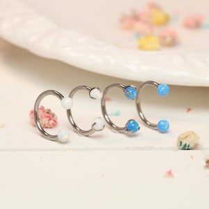 Puede incluir: Cuatro piezas de joyería corporal plateadas en forma de herradura con extremos de bola de ópalo y ópalo azul. Las joyas están dispuestas en fila sobre una superficie blanca, con un plato blanco y pequeñas decoraciones florales en el fondo.