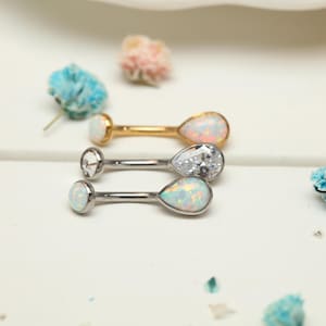 Pode incluir: Três piercings para o umbigo com detalhes em opala e cristal. Um é dourado, outro prateado com um cristal transparente e o terceiro prateado com uma lágrima de opala. As joias são exibidas em uma superfície branca.