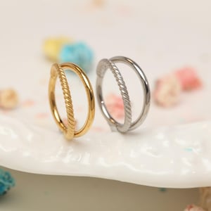 May include: Two hoop earrings, one gold-toned and one silver-toned, are displayed on a white surface. Each earring features a twisted rope design on one side and a smooth, rounded hoop. The earrings are set against a blurred background.