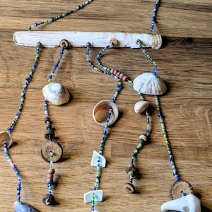 May include: A handmade wind chime with a driftwood base and strands of colourful beads. Shells, wooden beads, and small white shapes hang from the strands. The wind chime is displayed on a wooden surface.