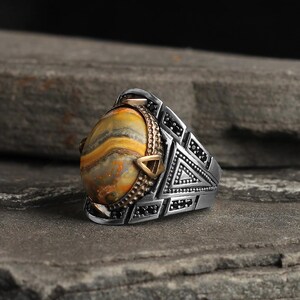 May include: A silver ring with an oval-shaped, yellow and brown stone. The ring has a geometric design with black accents and gold-colored details around the stone. The ring is set on a gray stone surface.
