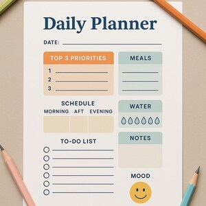 May include: A white daily planner with the title "Daily Planner" in navy blue. The planner includes sections for top priorities, meals, water intake, notes, and a to-do list. Colored pencils in green, orange, pink, and blue surround the planner.