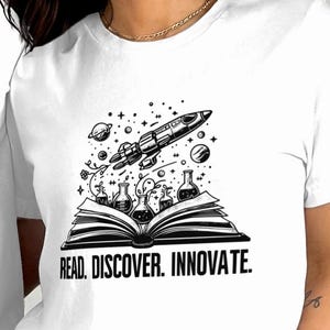 May include: White t-shirts featuring a black and white graphic of an open book with a rocket ship, planets, and beakers. The text "READ. DISCOVER. INNOVATE." is below the book.