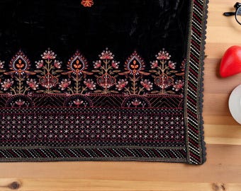 以下が含まれることがあります： 赤、金、白の精巧な花柄刺繍が施された黒い織物。 デザインは、様式化された花と幾何学模様の繰り返しパターンを特徴としています。 織物は木製の表面に置かれ、コーヒー、赤い唐辛子、眼鏡が添えられています。