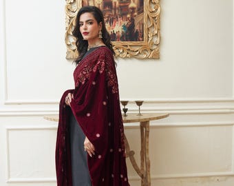 May include: A woman wearing a dark red velvet shawl with intricate embroidery over a gray outfit. The shawl drapes elegantly, showcasing detailed floral patterns. The background includes a framed painting and a small wooden table.