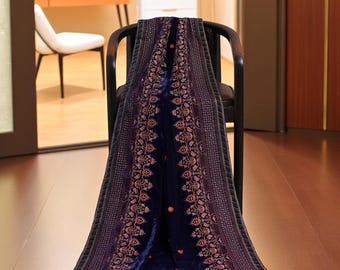 以下が含まれることがあります： ピンク、赤、オレンジの色合いで複雑な花の刺繍が施された、濃い青色のベルベットのショール。 ショールは黒い椅子に掛けられており、その詳細なデザインと豪華な質感を際立たせています。 背景にはマネキンに茶色のコートがあります。