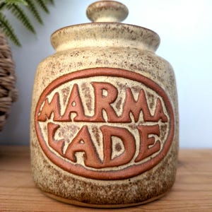 May include: A ceramic marmalade jar with a lid. The jar is beige with brown speckles and a brown oval with the word "MARMALADE" in raised letters. The lid has a small knob on top. A vintage kitchenware item.