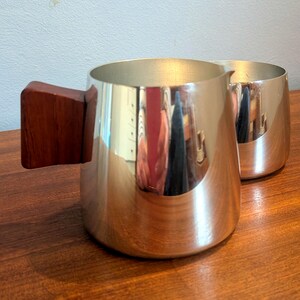 May include: Two silver-coloured pitchers with a reflective surface sit on a wooden table. One pitcher has a rectangular, dark brown wooden handle. The other pitcher is in the background. The pitchers are likely for serving beverages.