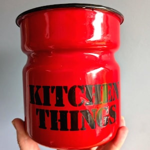 May include: A vibrant red enamel kitchen storage jar with a black rim. The words "KITCHEN THINGS" are printed in black. The jar has a cylindrical shape with a wider middle section, perfect for kitchen storage.