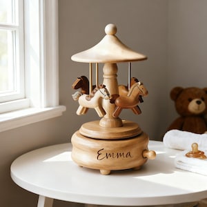 May include: A wooden carousel music box featuring three carved horses in varying shades of brown. The base is circular with a smooth finish and the name Emma is engraved in cursive script on the front. A small wooden knob is attached to the side.