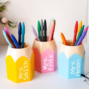 May include: Three pencil-shaped pen holders in yellow, pink, and blue, each with a teacher's name printed on the side. The holders are filled with pens, pencils, and a comb. The pencil holders are on a white surface.