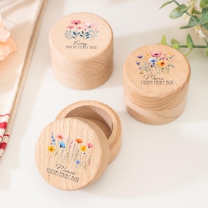 Może przedstawiać: Trzy drewniane pudełka na zęby wróżki z kwiatowymi wzorami. Każde pudełko ma pokrywkę z innym akwarelowym układem kwiatów i imieniem. Tekst "TOOTH FAIRY BOX" jest wydrukowany pod imieniem na każdym pudełku.
