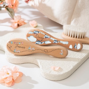 Puede incluir: Cepillos para el cabello de bebé de madera con cerdas suaves. Dos cepillos están decorados con ilustraciones coloridas y los nombres "James" y "Daisy". Los cepillos tienen un acabado de madera clara y están sobre una superficie blanca.